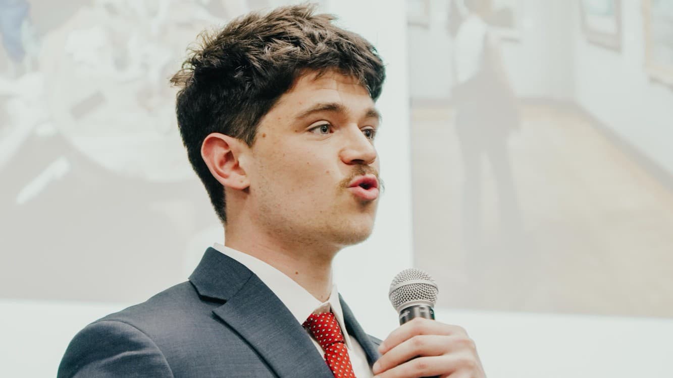 Judson Dunne mid-sentence with a microphone, in a navy suit and red tie
