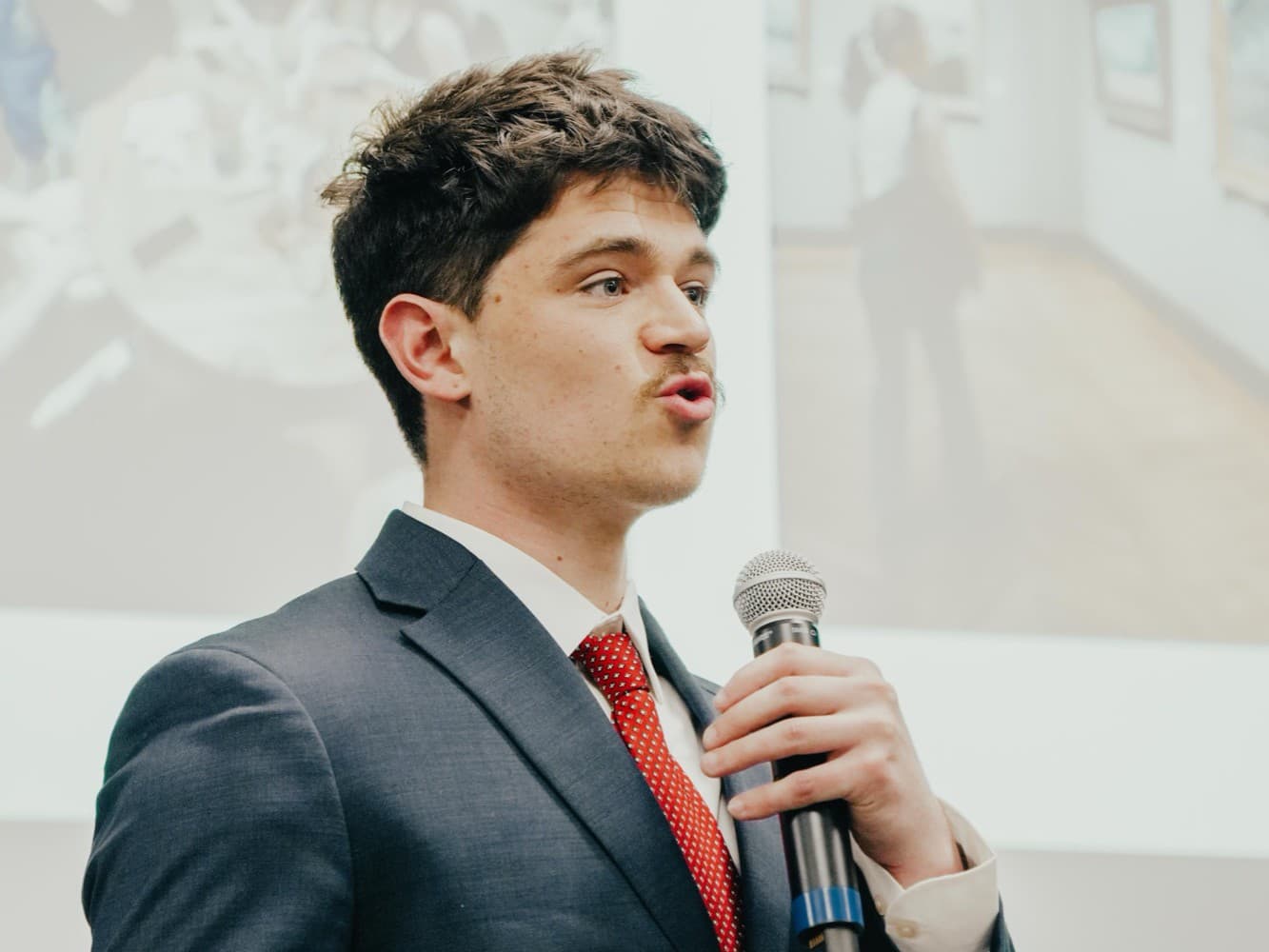 Judson Dunne in a navy suit and red tie, presenting with a microphone