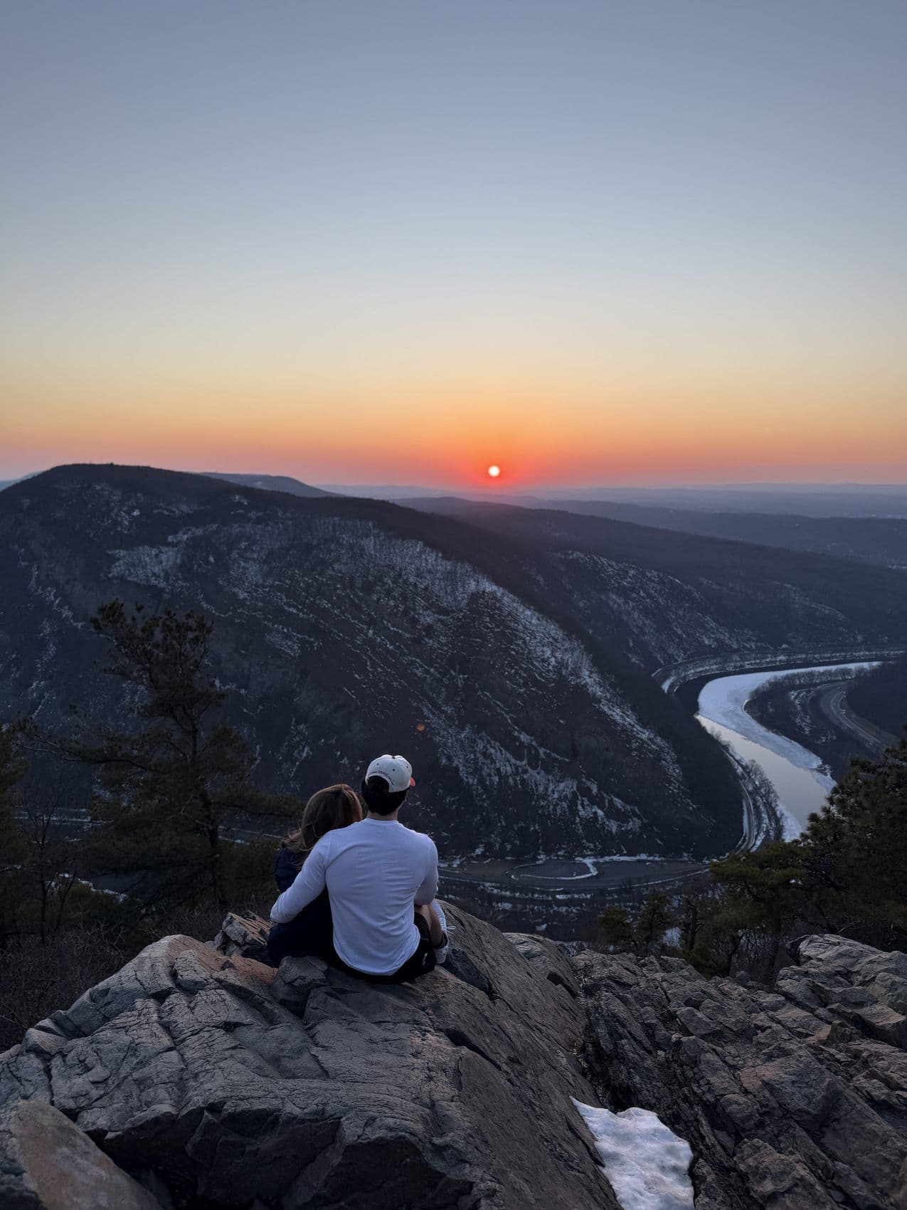 Two people on a cliff at sunset above a river valley